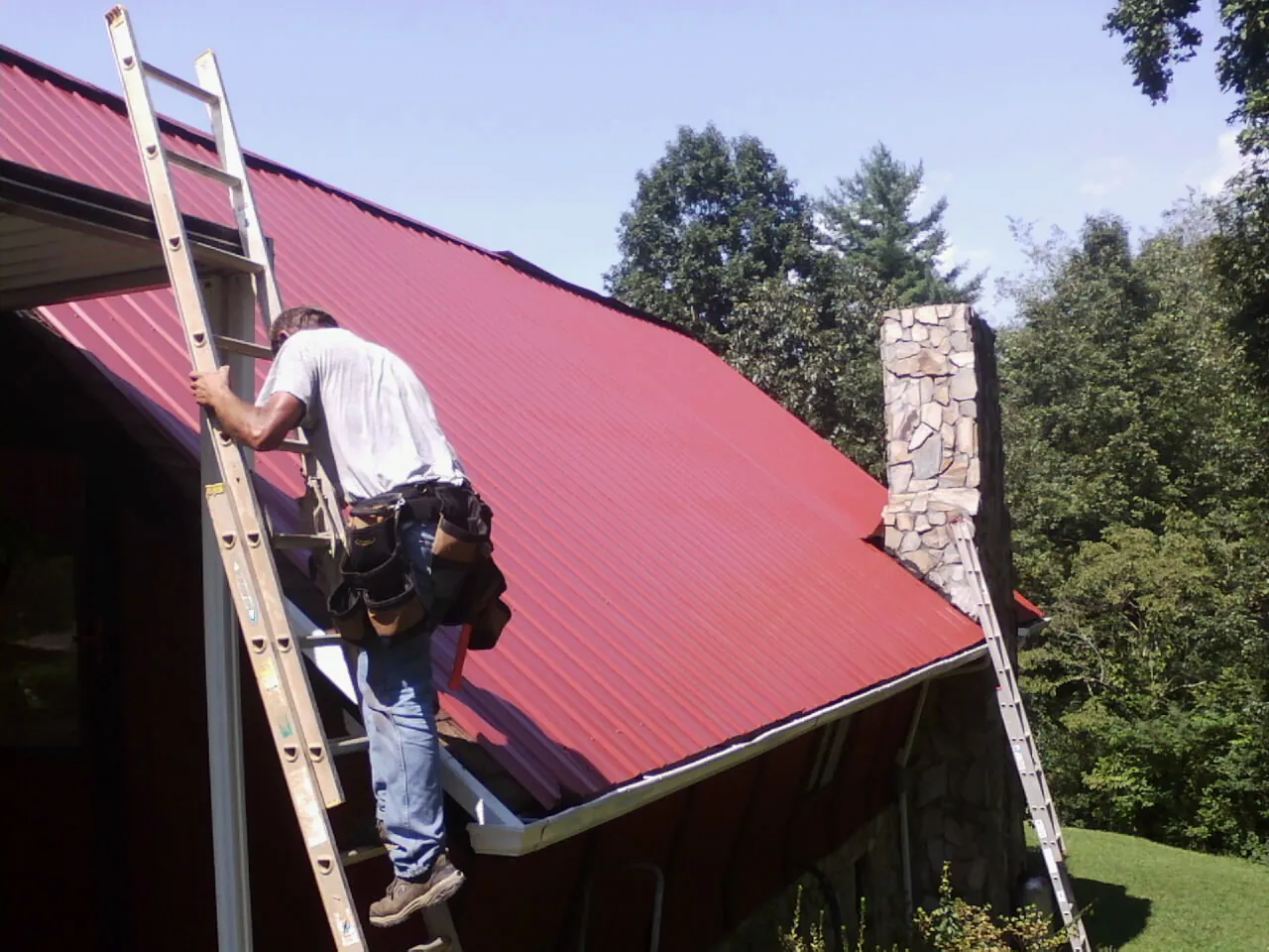 Roofer on a ladder beside a red metal roof and stone chimney — Soffit & Fascia Repair by Roofing Brothers in Thomasville